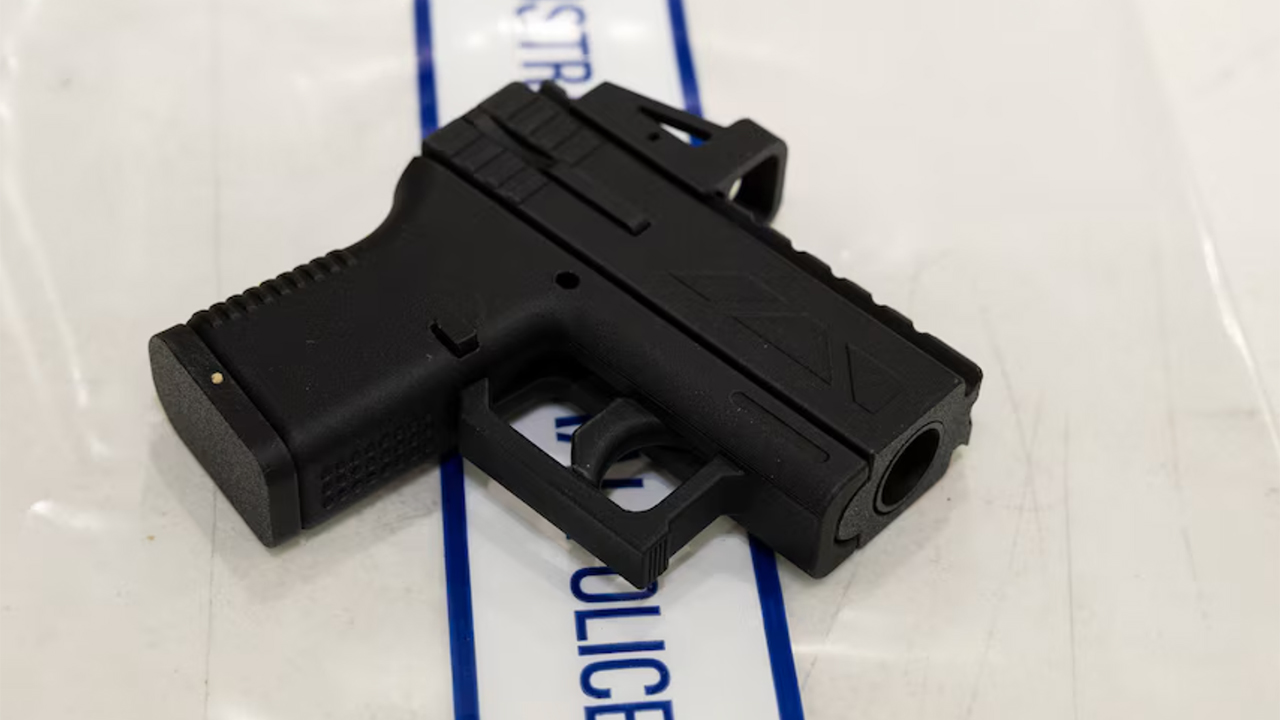 Black semi-automatic pistol lying on a white surface with a blue stripe in the background, viewed from the side.