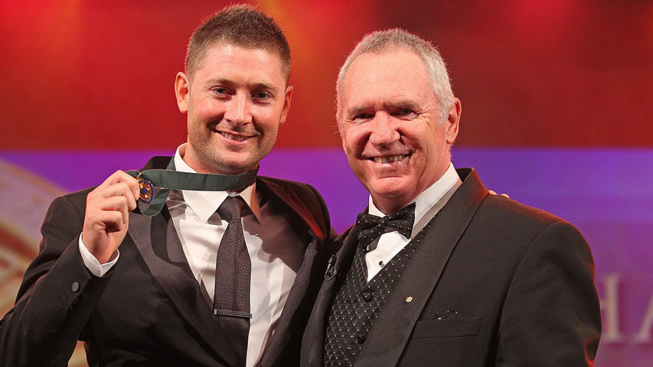 Two men in tuxedos smiling on stage; the younger man holds a medal on a ribbon toward the camera.