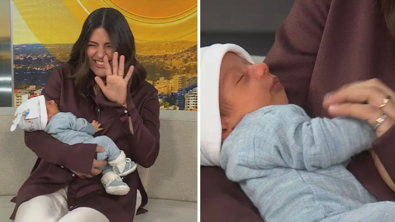 Woman in a burgundy coat cradles a newborn wearing light blue clothing, smiling and waving at the camera with the baby resting in her arms.