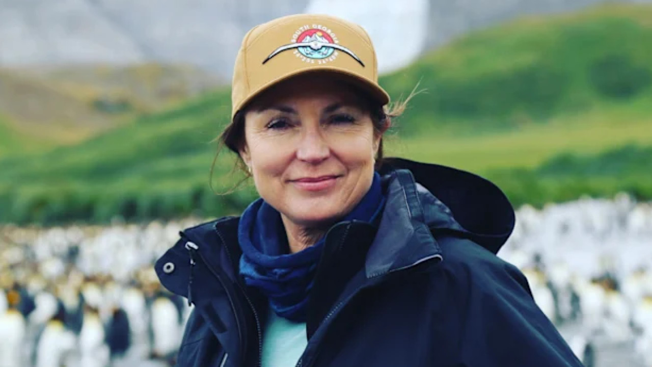 Smiling woman in a tan cap and navy jacket with a blue scarf, outdoors with a colony of penguins in the background.