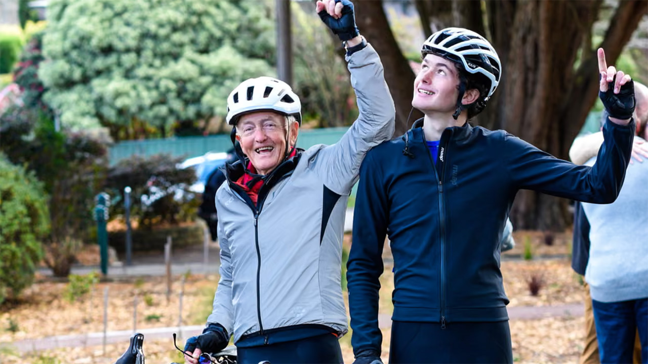 Two cyclists wearing helmets and jackets celebrate with raised arms in a park after a ride, smiling at the camera beside their bikes.