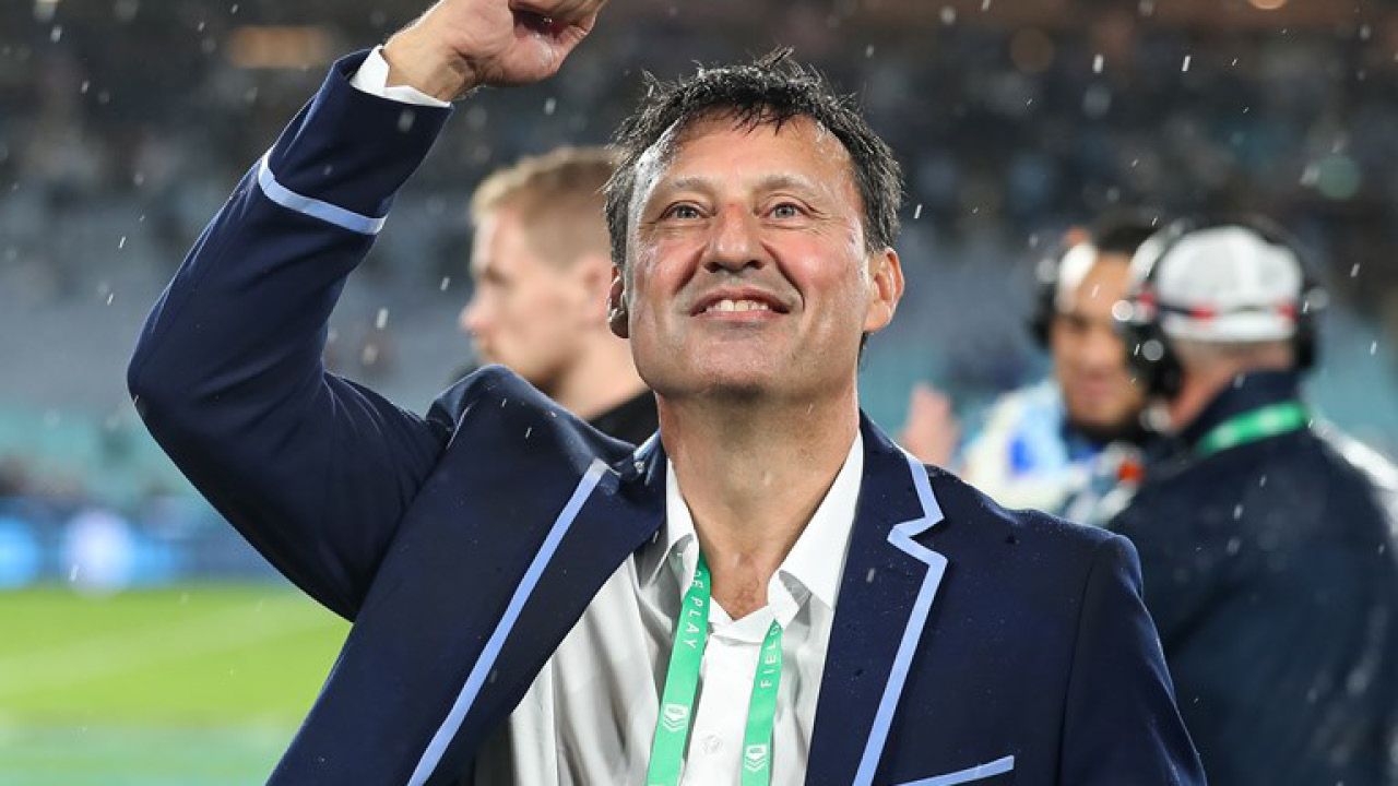Man in a navy blazer with light blue trim raises his arm in celebration on a snowy stadium field.
