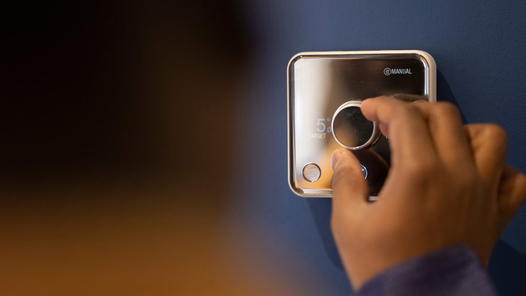 Close-up of a hand adjusting a thermostat knob on a blue wall; shows 5° target and manual mode label.