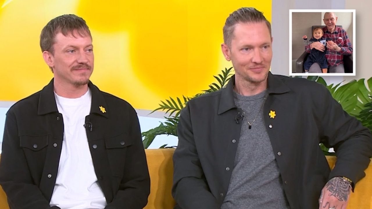 Two men sit on a yellow couch in a TV studio, wearing black jackets with yellow daffodil pins; a small inset photo shows a grandfather with a baby.