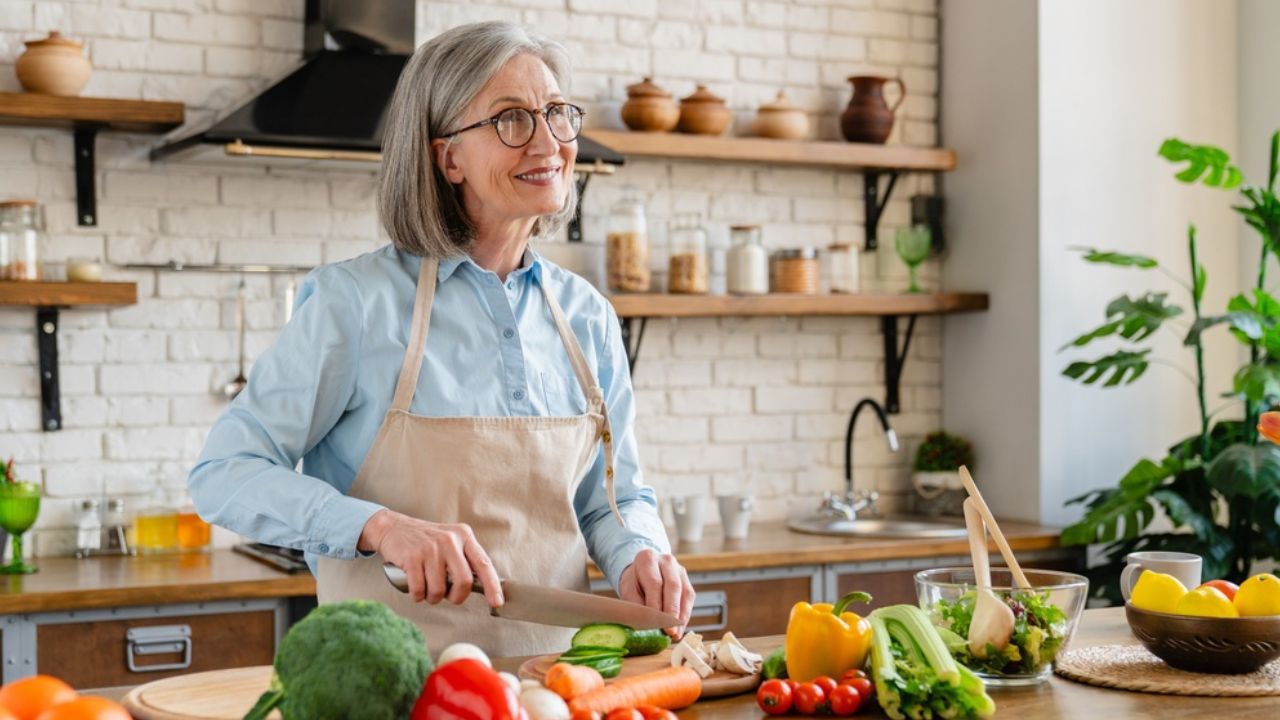 Older woman with gray hair and glasses wearing a beige apron chops vegetables on a wooden kitchen counter, with peppers, cucumber, and broccoli nearby in a bright, plant-filled kitchen