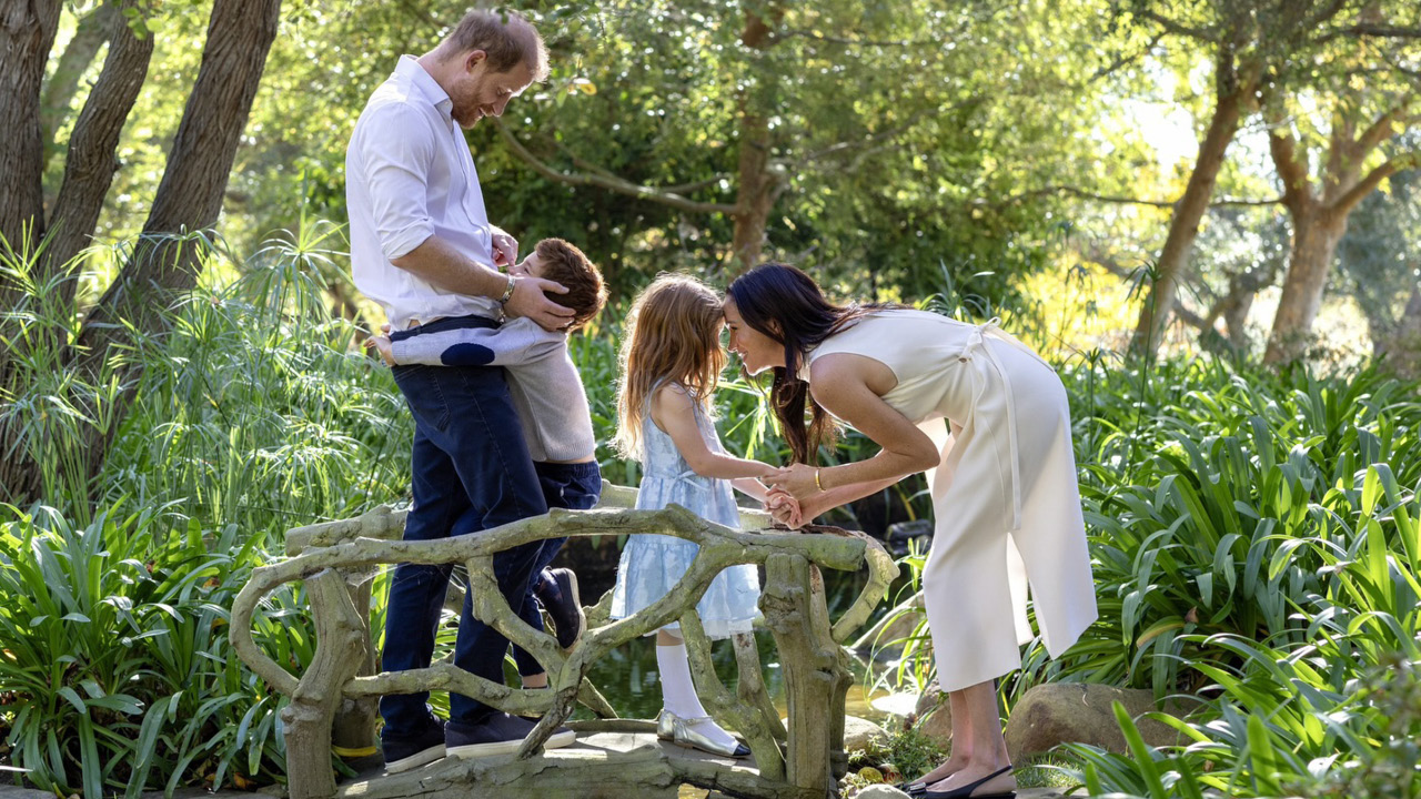 Family outdoors in a green garden; father holds a baby while mother leans to speak with their daughter on a wooden bridge.