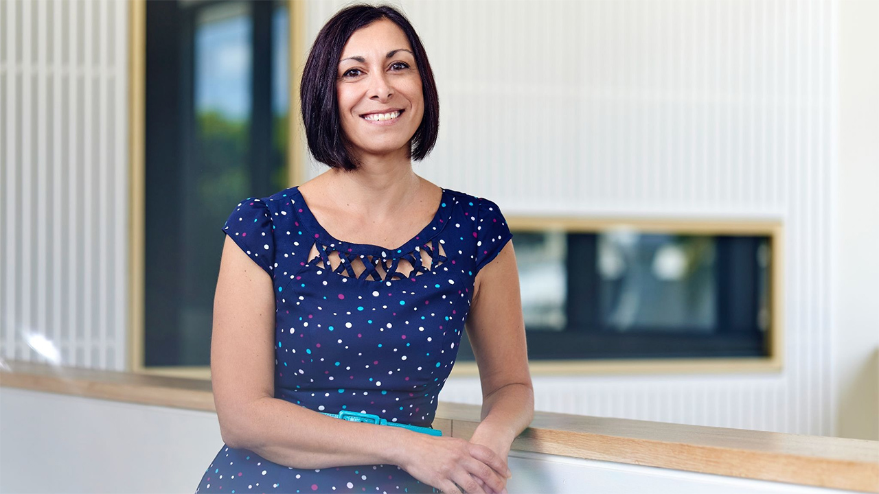 Portrait of a smiling woman with short dark hair, wearing a navy polka-dot dress, leaning on a wooden railing inside a modern building.