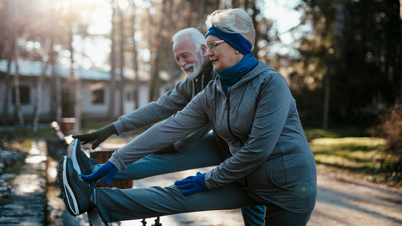 Two elderly adults in athletic wear stretch their legs on stationary bikes outdoors in a sunlit park.