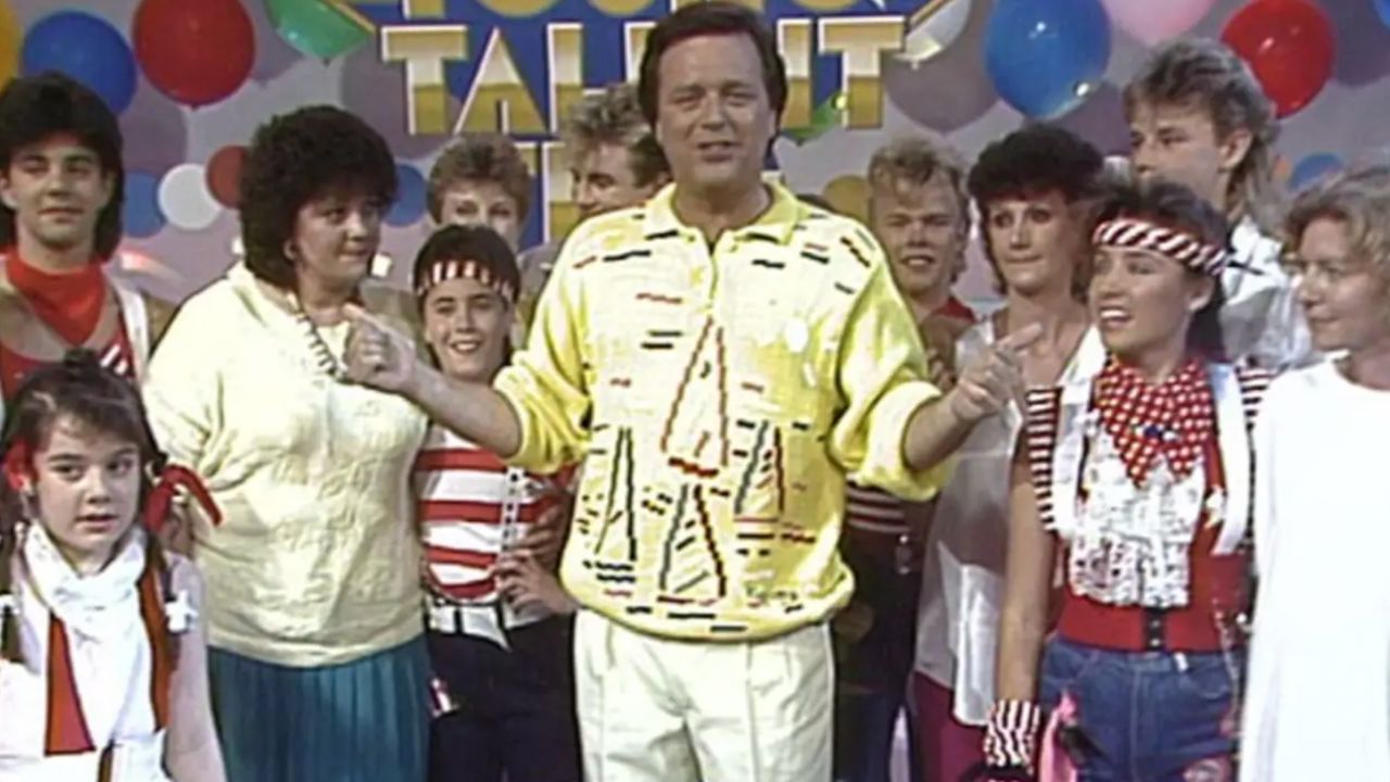 Host in a yellow patterned shirt stands center among a group of performers in 1980s outfits on a colorful stage with balloons and a 'Talent' sign in the background.