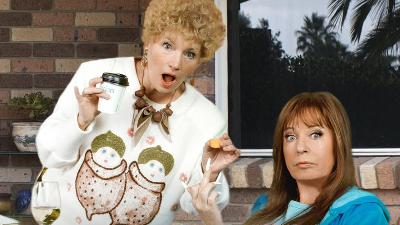 Two women on a brick porch; one with curly blond hair in a white sweater shows a small sponge and a nail tool, the other woman with brown hair looks surprised nearby.