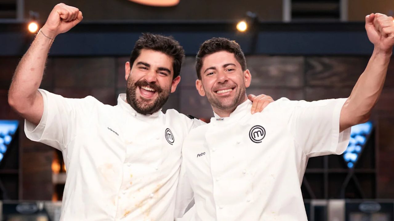 Two male chefs in white jackets celebrate with arms around each other, smiling broadly in a kitchen set.