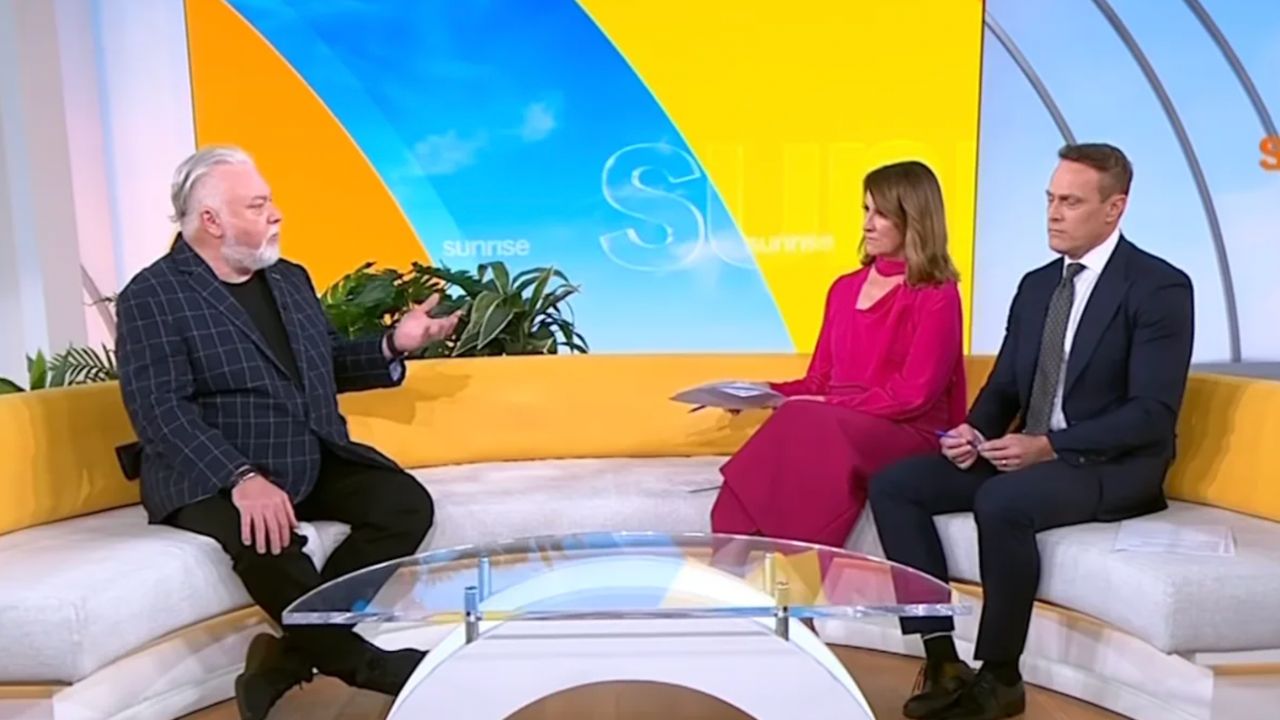 Three people on a bright morning TV set with a yellow curved couch; the guest speaks while two hosts listen.