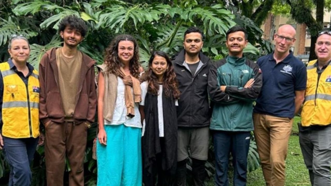 Group of eight diverse people standing together outdoors in front of large tropical plants, smiling at the camera.