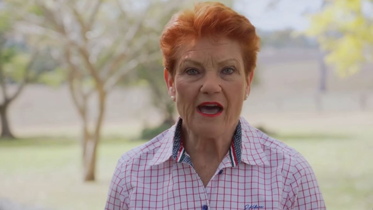 Older woman with short red hair speaking outdoors, wearing a white and pink checkered shirt, with trees in the background.