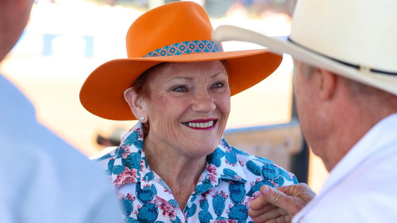 Smiling older woman in an orange wide-brim hat and floral shirt chats with a man outdoors.