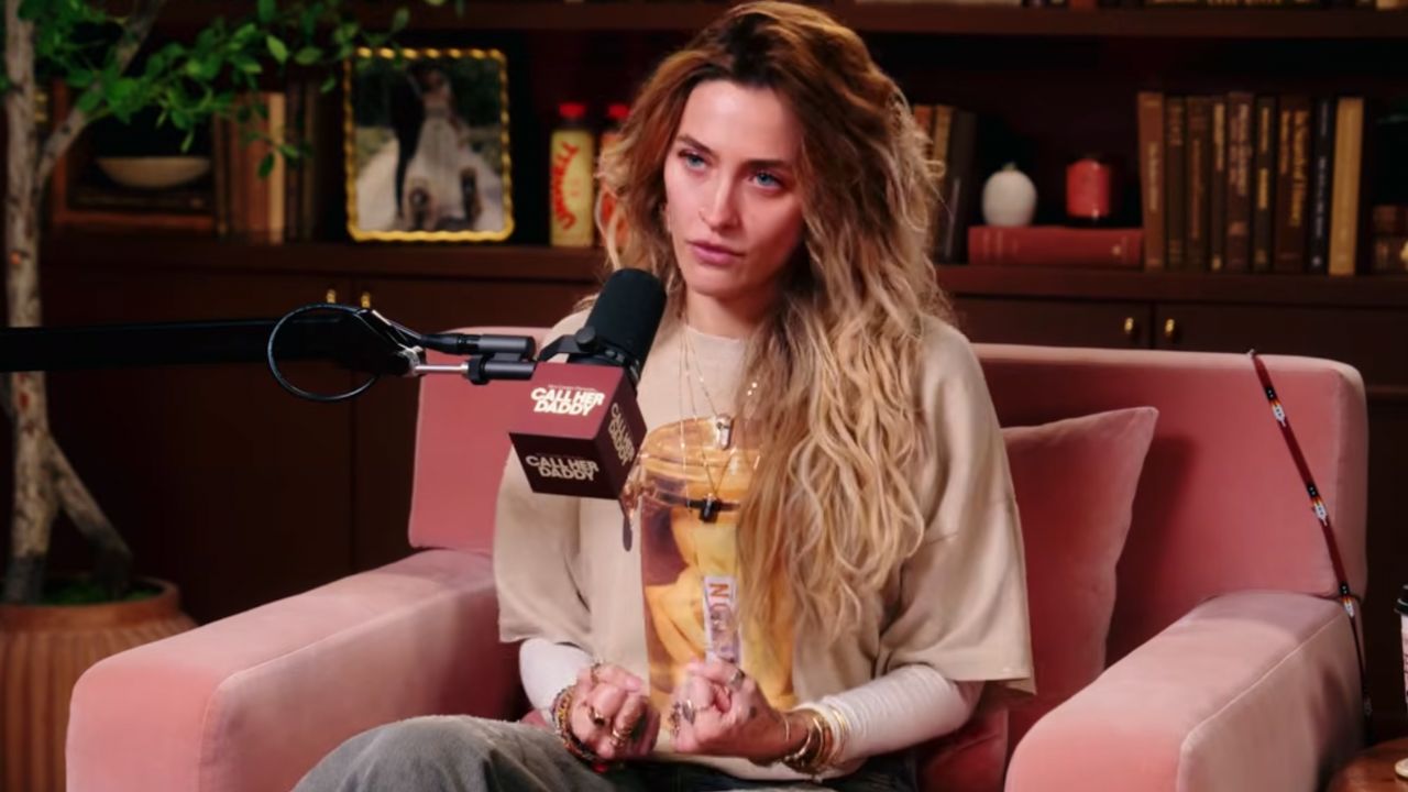 A woman guest seated on a pink sofa, speaking into a mounted podcast microphone during an interview in a cozy studio set.