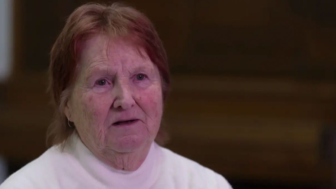 Close-up of an elderly woman with short reddish hair wearing a white top, looking slightly to the left indoors.