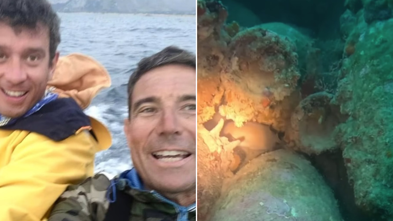Two men on a boat taking a selfie, wearing jackets, open water and a distant shoreline in the background.