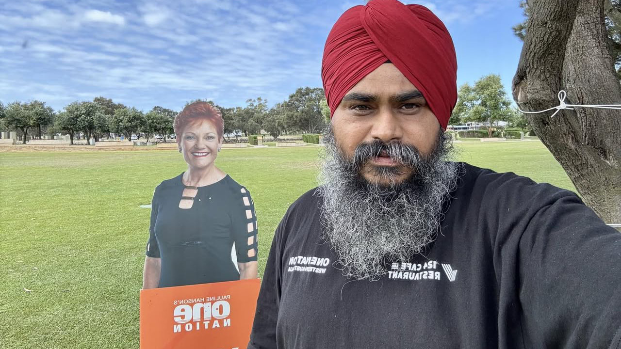 Man with a red turban and long beard takes a selfie beside a life-size cardboard cutout of a smiling woman holding an orange sign that reads 'One Nation' in a grassy park area.
