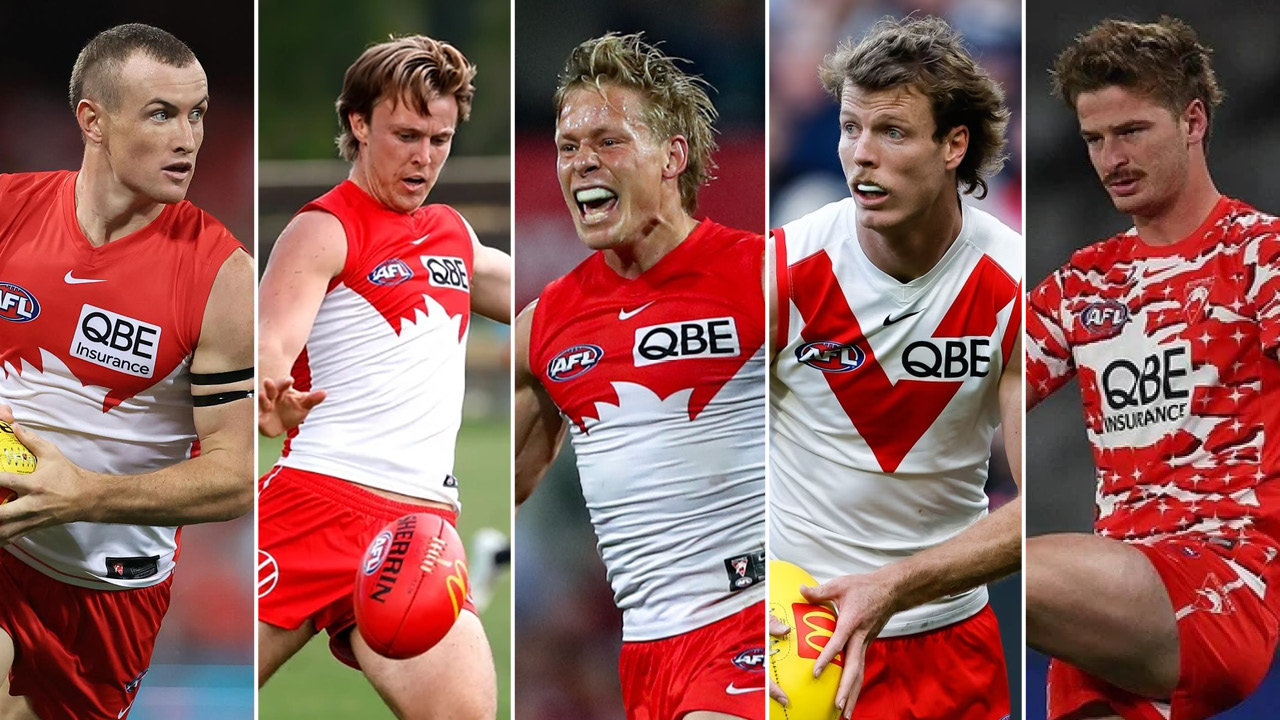 AFL player in red and white jersey holding a football, looking to the left. Sponsor patch QBE Insurance visible on chest.