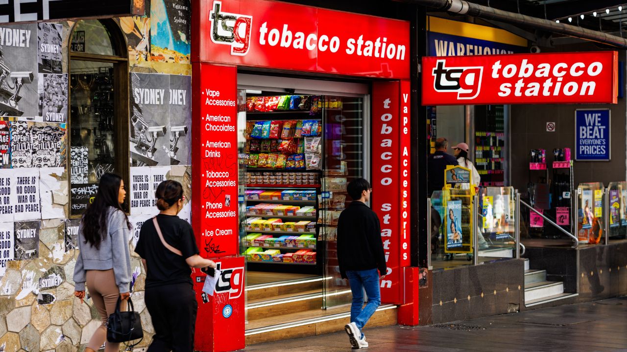 Red storefront of a tobacco station shop with snacks visible inside and pedestrians passing by on the street.
