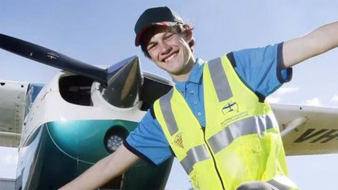 Young man in a yellow safety vest and blue shirt posing with a small airplane and its propeller.