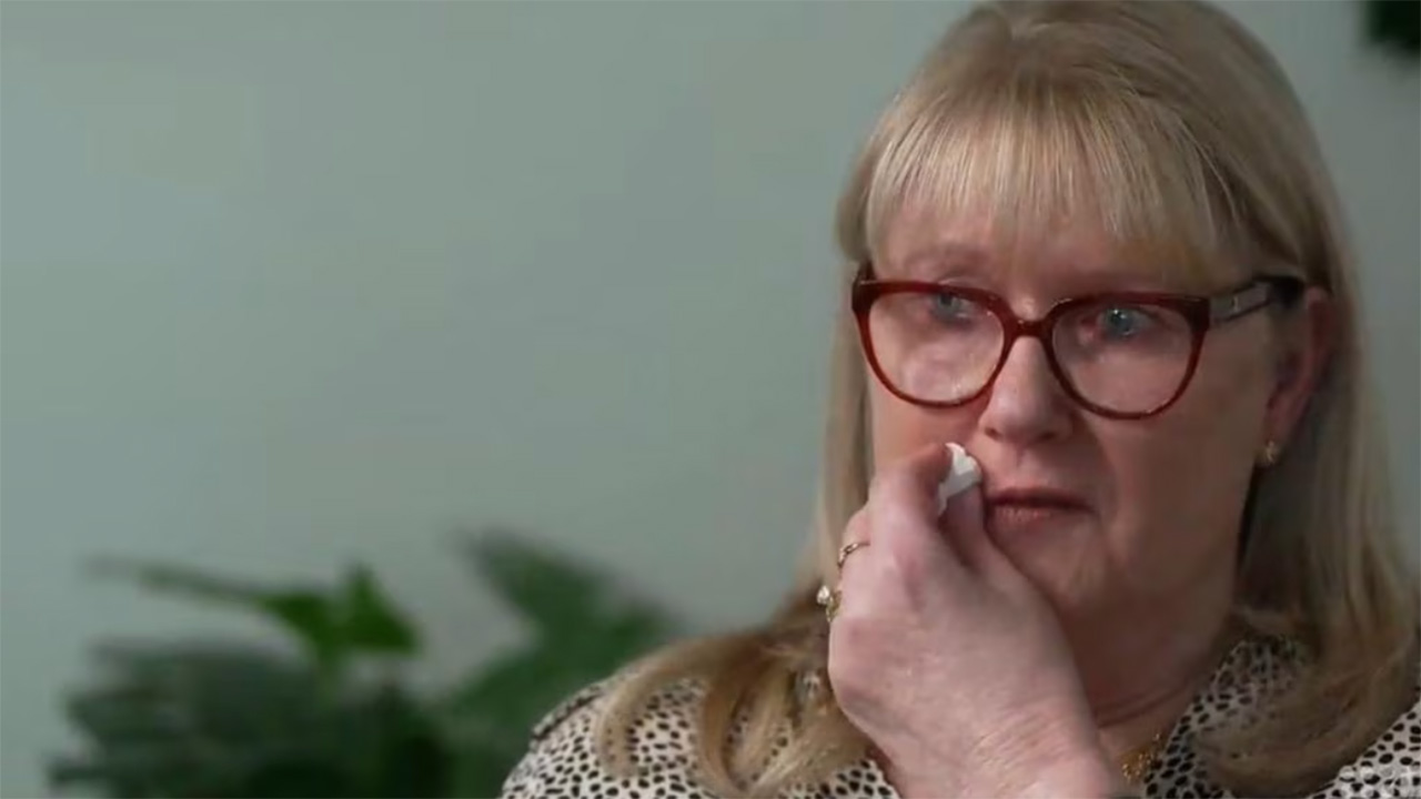Older woman with red glasses touching her nose with a tissue or cotton, wearing a patterned blouse