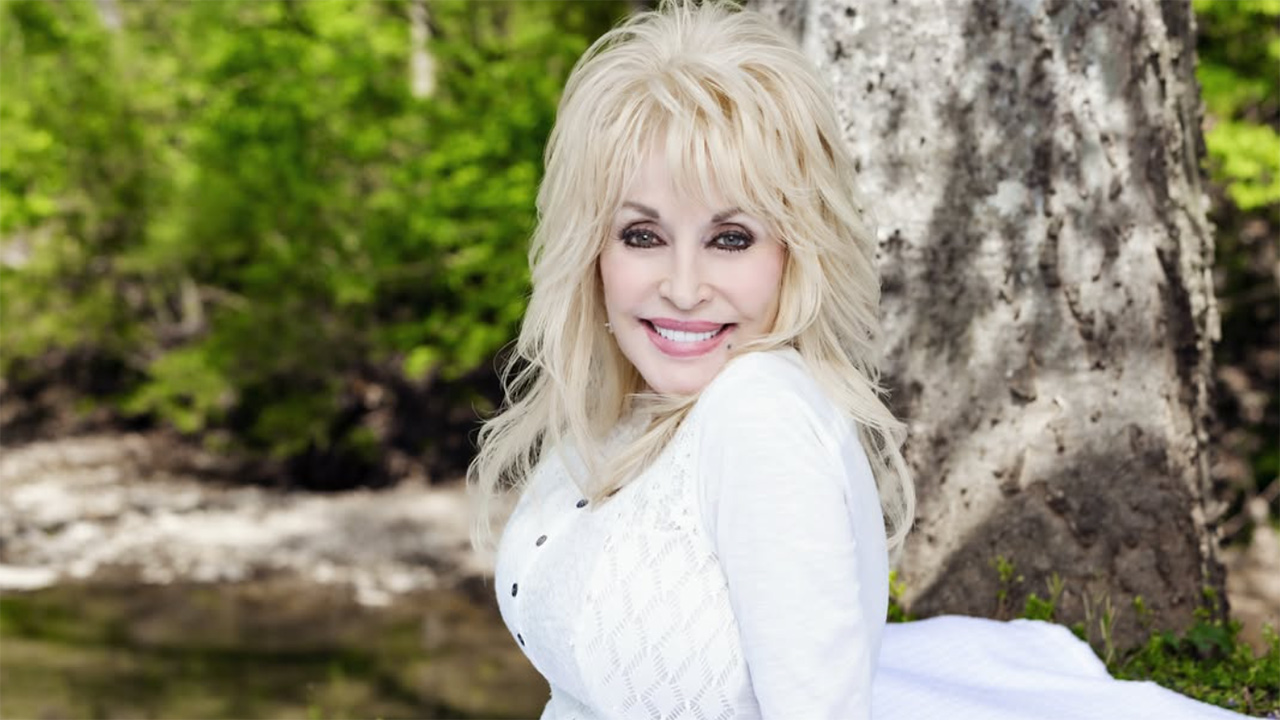 Smiling blonde woman in white top posing outdoors by a tree near a lake.