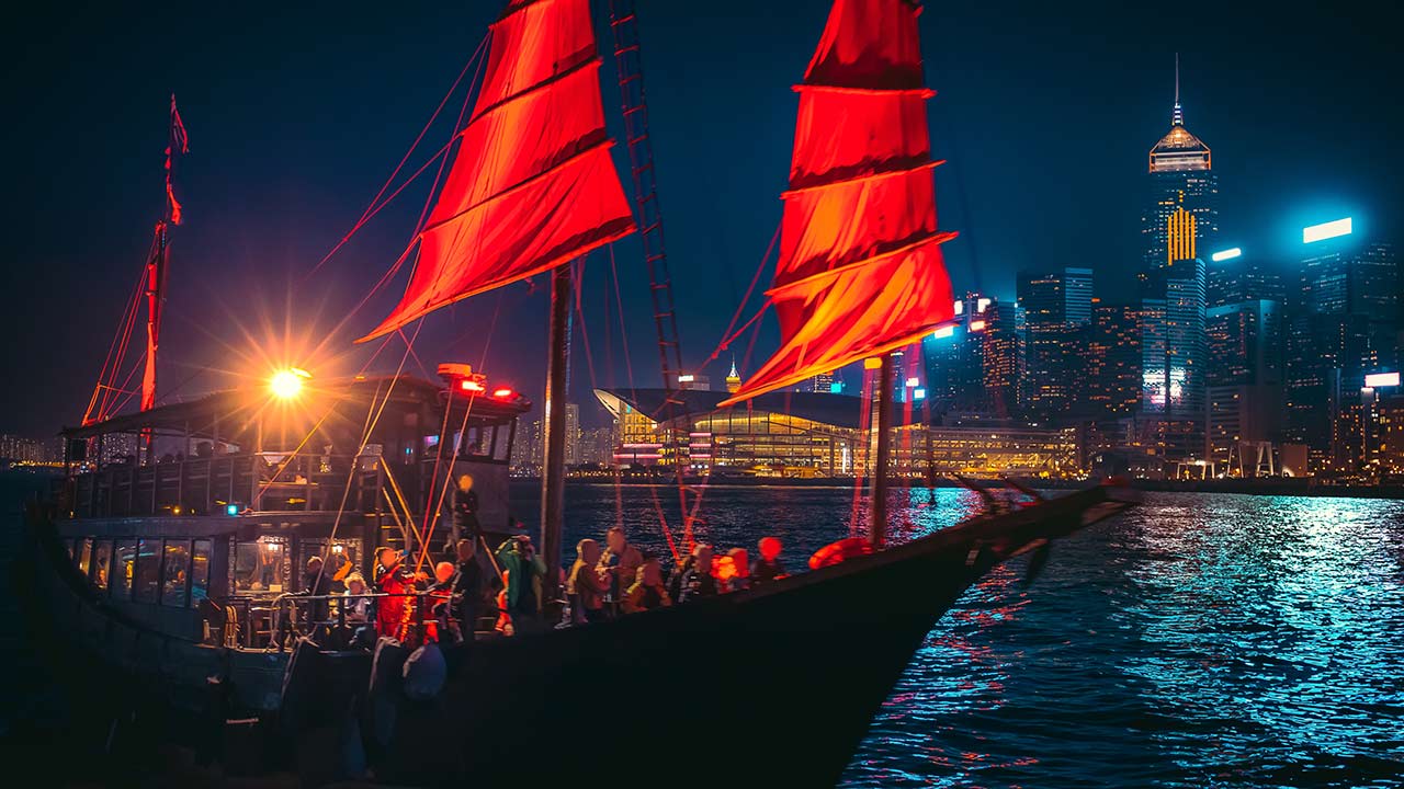 Nighttime harbor scene with a sailing boat and bright red sails against a lit city skyline across the water.