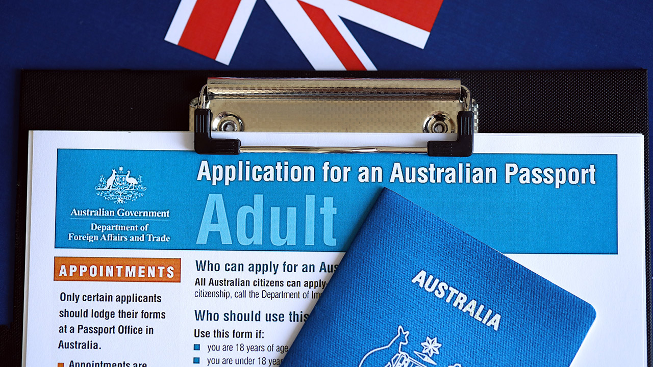 Australian passport application form on a clipboard with a blue passport cover partially overlapping it on a desk with a flag backdrop.