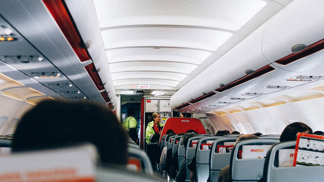 Inside a commercial airplane cabin with passengers seated and a flight attendant standing in the aisle near the rear exit sign.