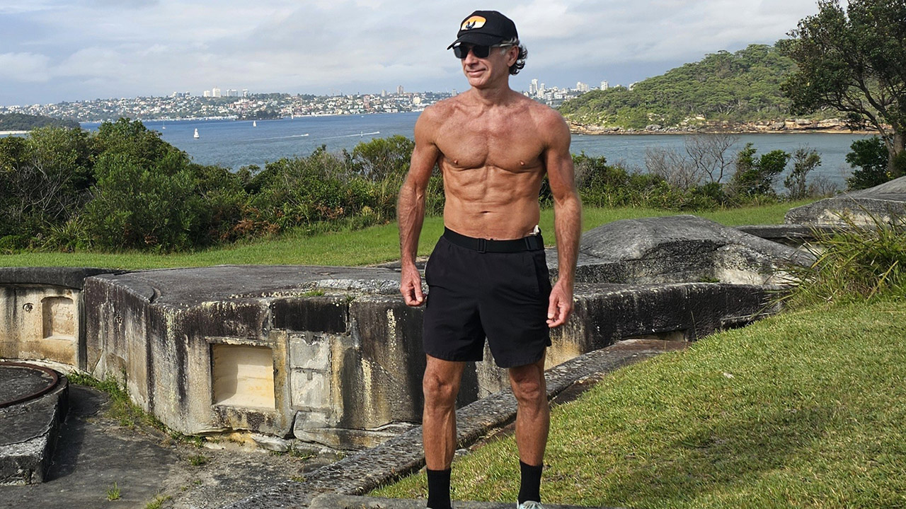 Muscular man in black shorts, cap, and sunglasses stands shirtless on a concrete seawall with water and cityscape in the background.