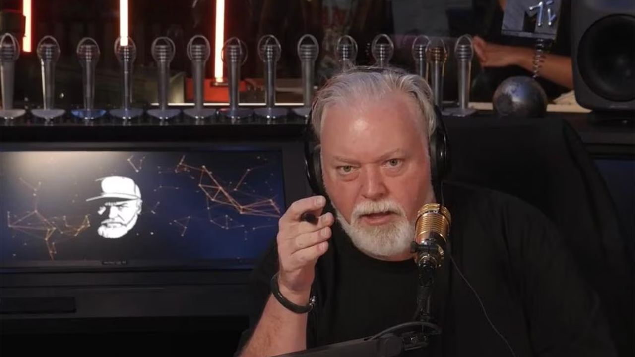 Radio host with white beard and headphones speaks into a gold microphone, pointing at the camera while on air.