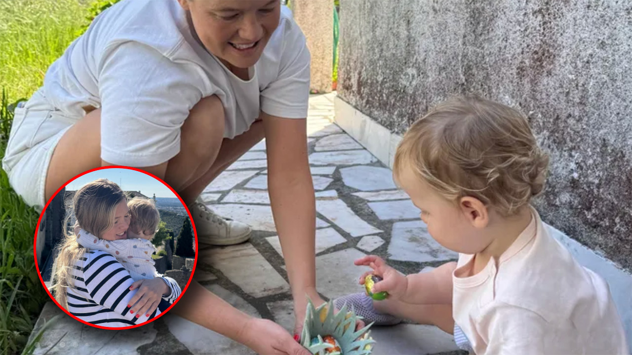 Adult woman crouching and smiling at a toddler who holds a colorful egg on a stone patio outdoors.