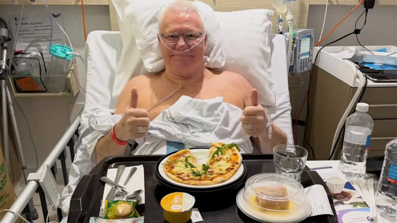 Man in a hospital bed giving a thumbs-up with an oxygen mask, a pizza on a tray in front, and a water bottle nearby.