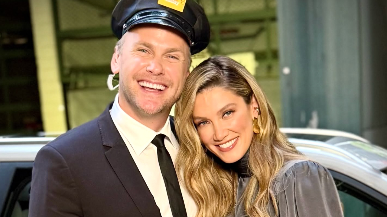 Smiling couple posing outdoors beside a car; man wears a dark suit and captain's hat, woman in a gray outfit.