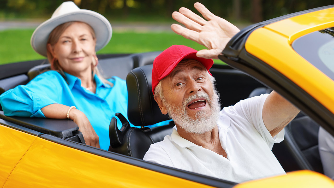 Older man with a white beard driving a yellow convertible, waving at the camera with a smile; woman sits beside him.