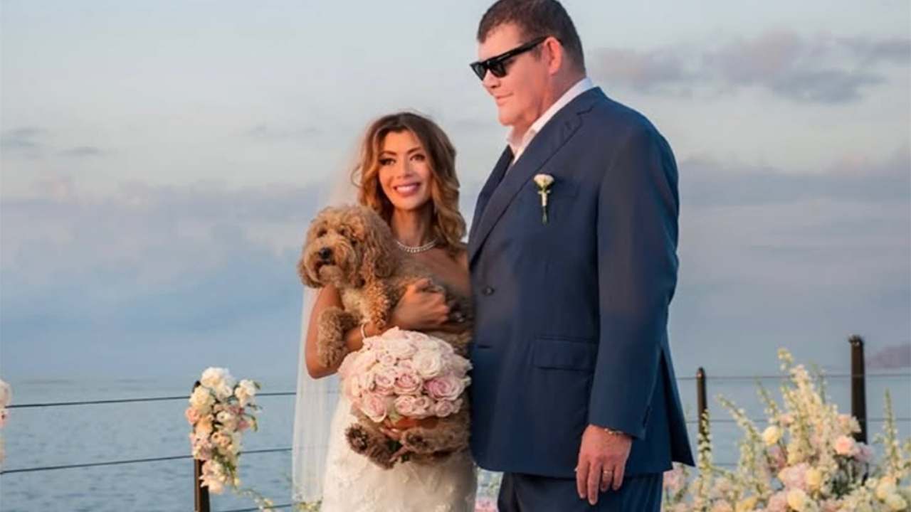 Bride in a white gown holds a pink rose bouquet and a small brown poodle, standing beside a groom in a blue suit by the water.