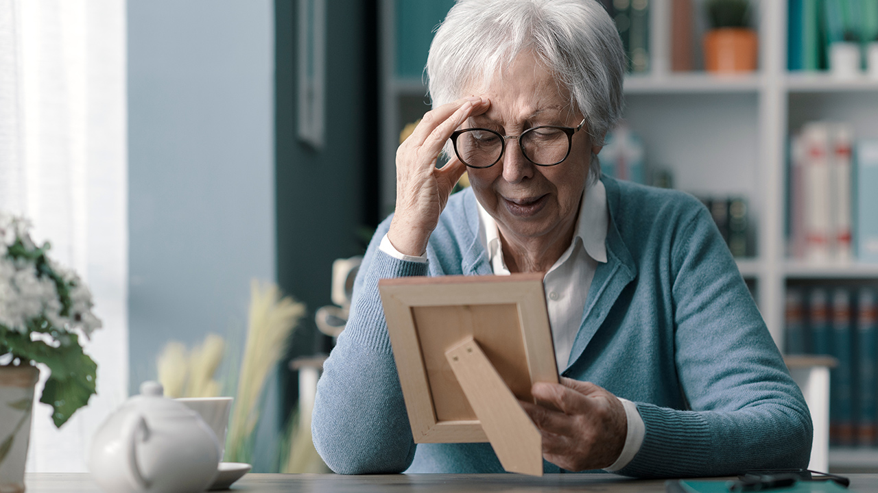 Older woman with short gray hair and glasses, holding a wooden photo frame and looking at it pensively at a desk in a cozy room