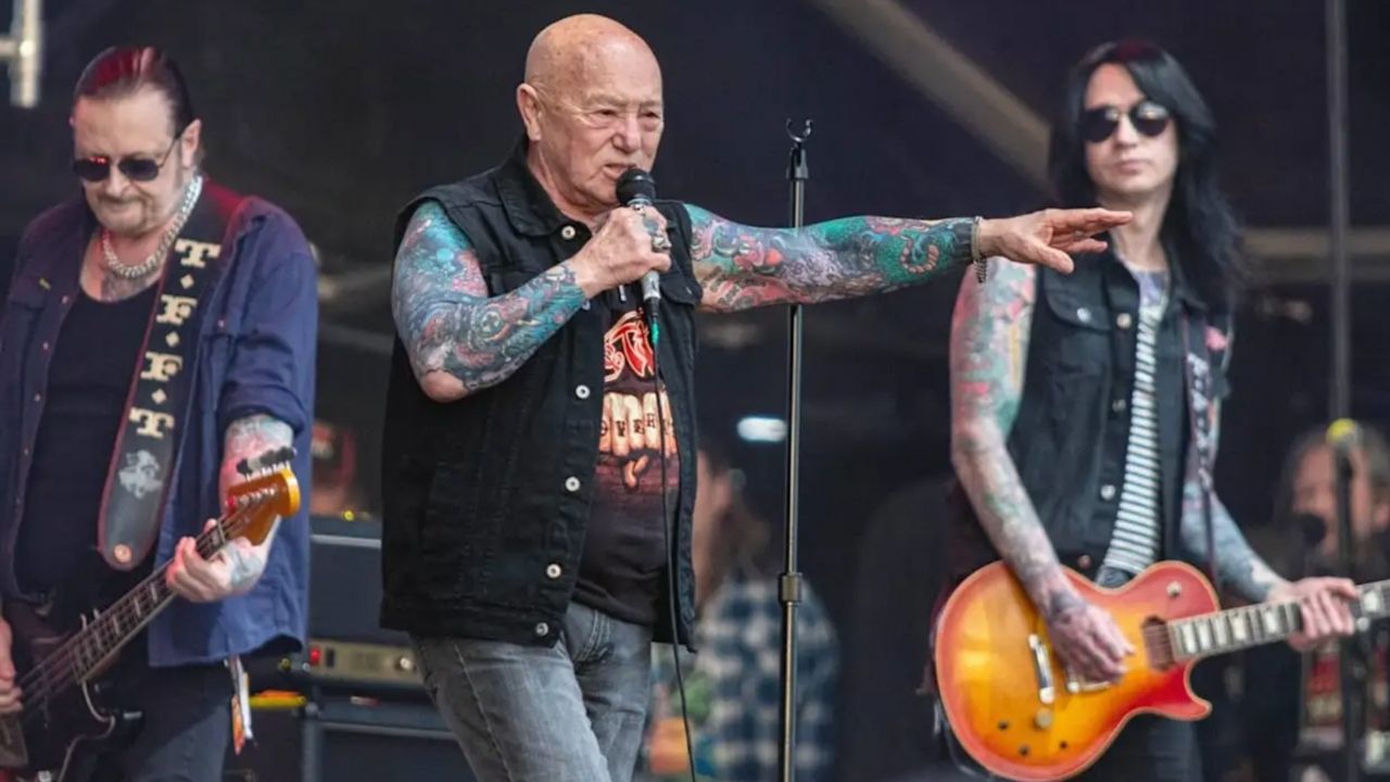 Lead singer with tattooed arms sings into a microphone on stage, flanked by two guitarists playing guitars on a rock show.